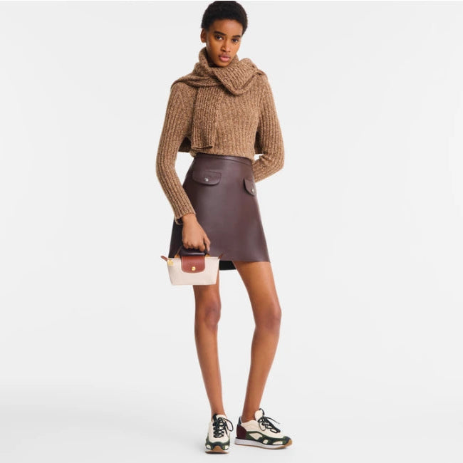 A model stands against a light gray studio background wearing a chunky knit cropped sweater with a high neck in camel brown, a dark brown faux-leather mini skirt, black and white sneakers, and holding a small cream and brown handbag.